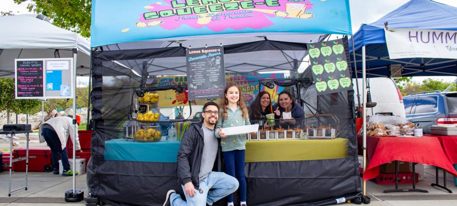 The smiling owners of Lemon Squeeze-E standing in front of their colorful booth at the Dublin Farmers' Market. A female child poses with them, extending a tray of stickers in her arms.