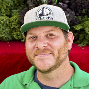 A photo of Dimitri Hagnere, a smiling bearded man wearing a hat sitting in front of farmers' market produce