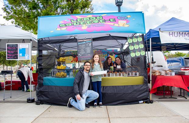 The smiling owners of Lemon Squeeze-E standing in front of their colorful booth at the Dublin Farmers' Market. A female child poses with them, extending a tray of stickers in her arms.