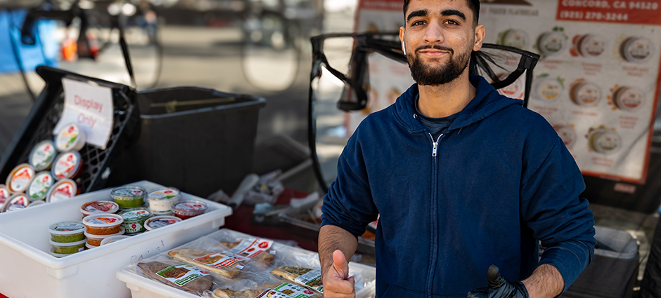 A seller standing in front of dips at the Bolani booth giving two thumbs up